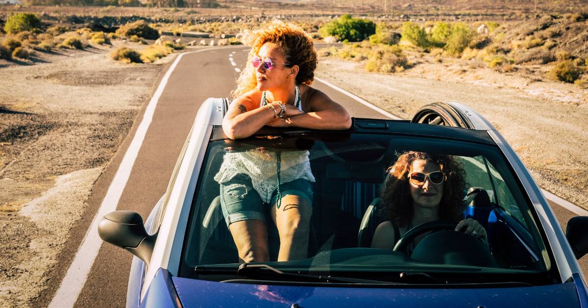 Two friends on a roadtrip in a convertible, one driving and the other sat on top of the passenger seat.