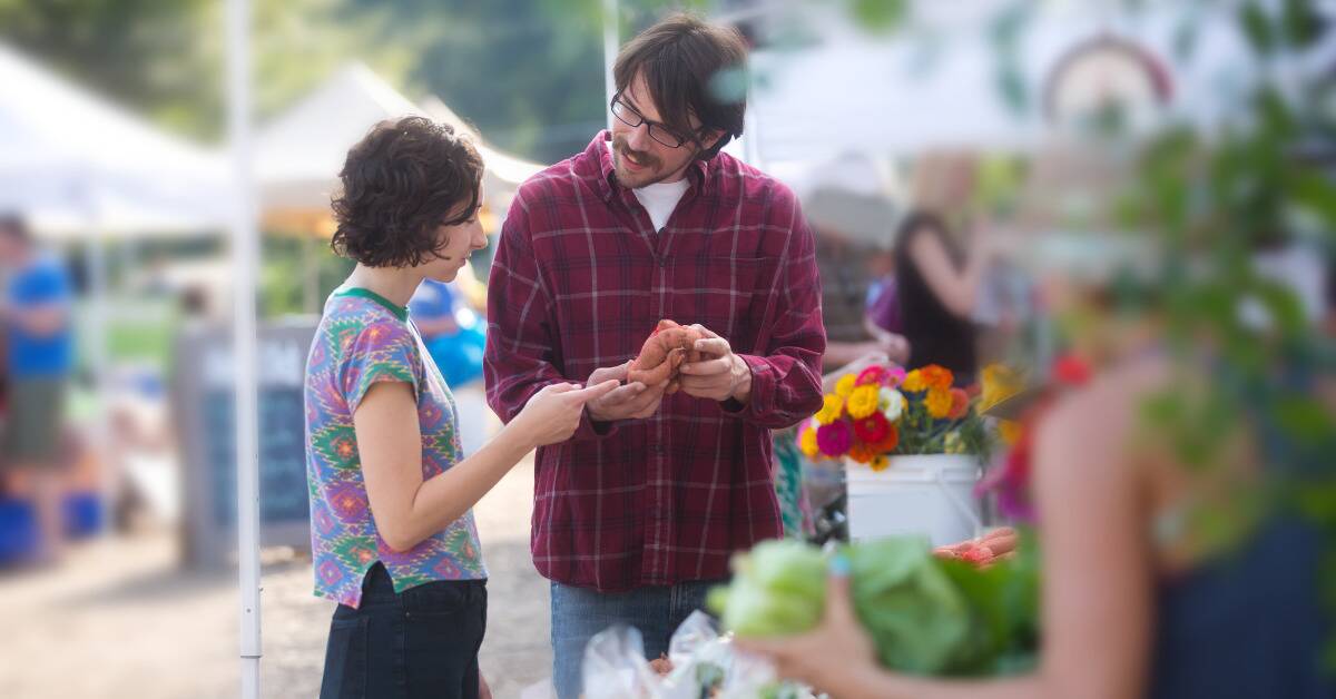 A couple at a farmer's market, the man showing the woman a sweet potato.