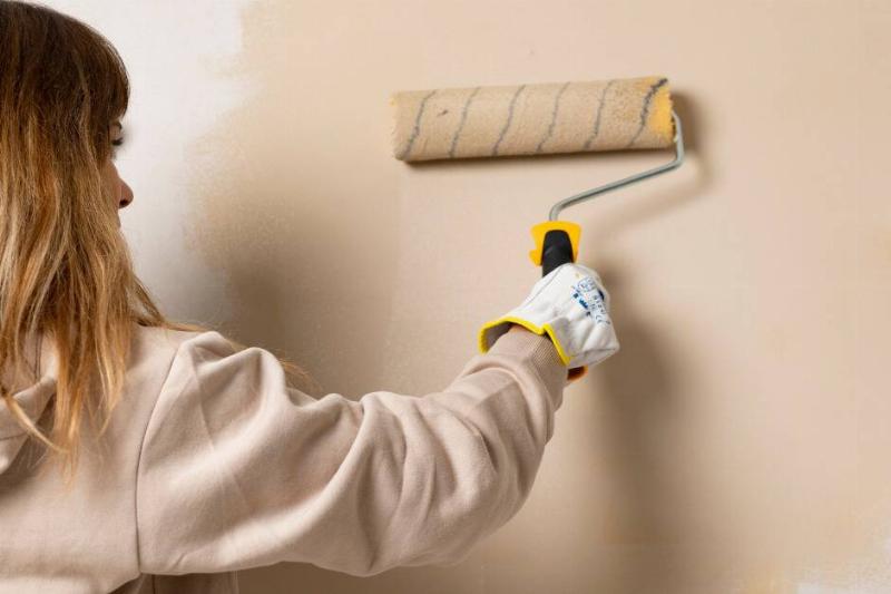 A woman using a paint roller to paint her wall beige.