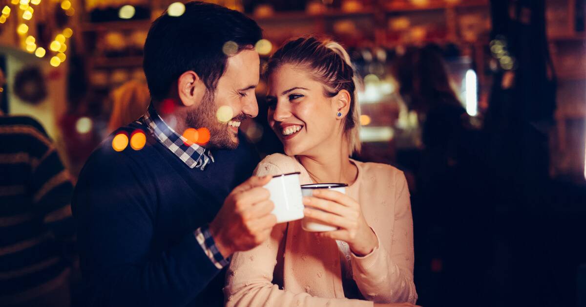 Two people cheers-ing mugs while on a date, sitting very close to one another.