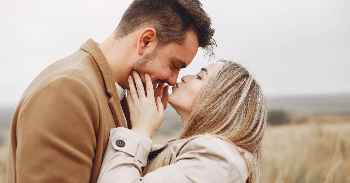 A couple standing close to each other, the man tilting his face down so the woman can gently kiss his nose.