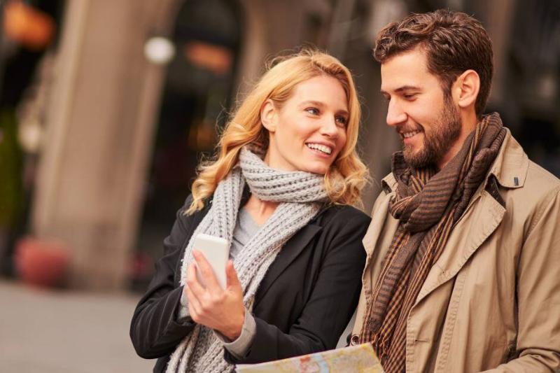 A couple walking outside, one holding a map and the other pointing to their phone, smiling at one another.
