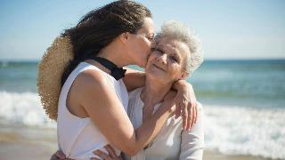 A mother and daughter on the beach hugging, the daughter kissing her mother's cheek.