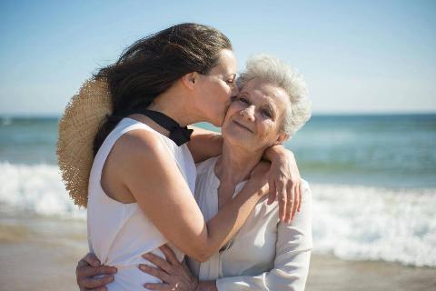 A mother and daughter on the beach hugging, the daughter kissing her mother's cheek.