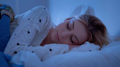A woman sleeping on her side in bed, hand under her head, in soft blue lighting.