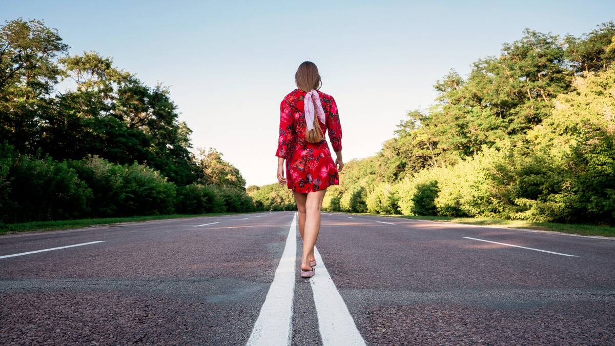 A woman in a bright red dress walks away from the camera on the center line of a two lane road.