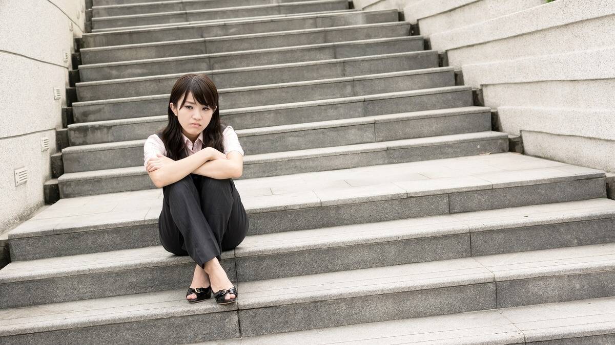 A woman sitting with her knees drawn up on a set of concrete stairs outside, looking discontented or even angry.