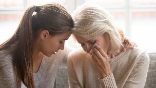 A mother and daughter sitting side by side, the daughter comforting the mother as she cries, the mother wiping away a tear off her cheek.