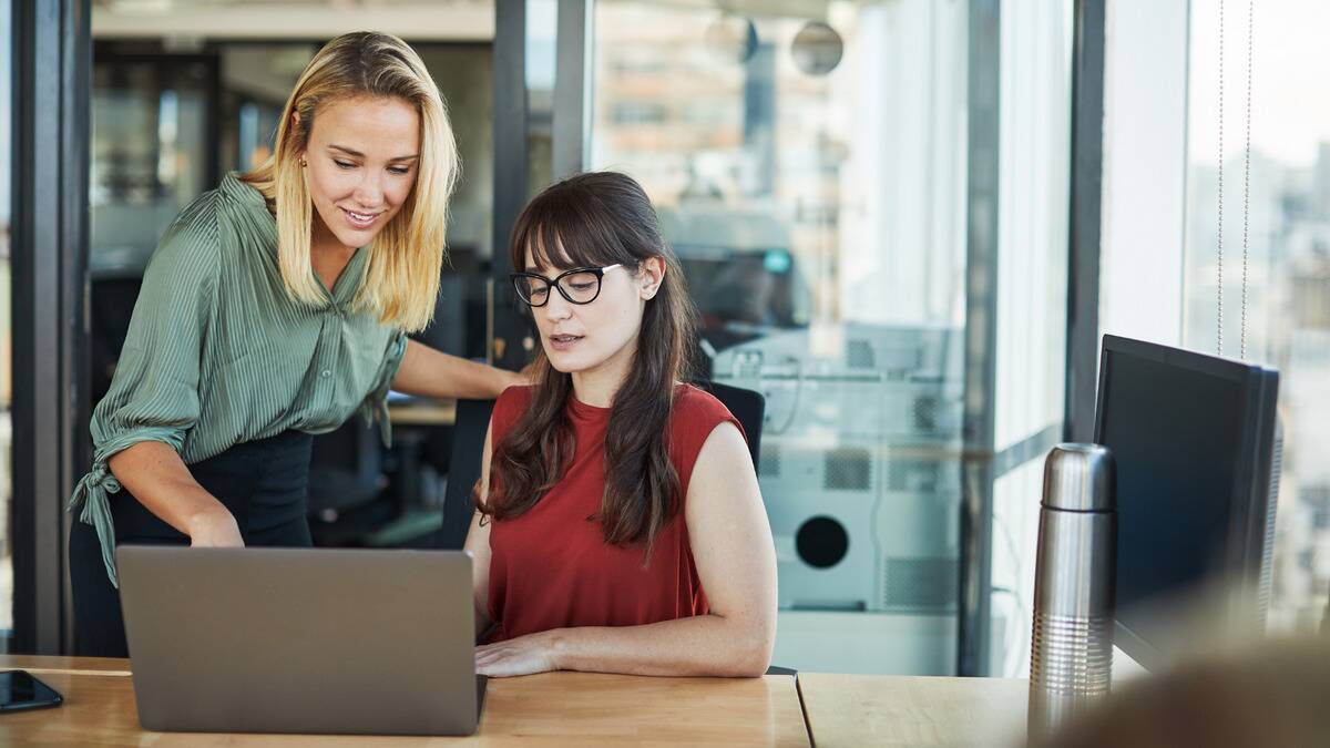 Two women looking at a laptop, one sitting and one standing pointing at something on screen.