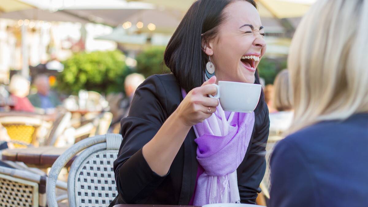 A woman laughing as she holds a mug, sitting outside with a friend.