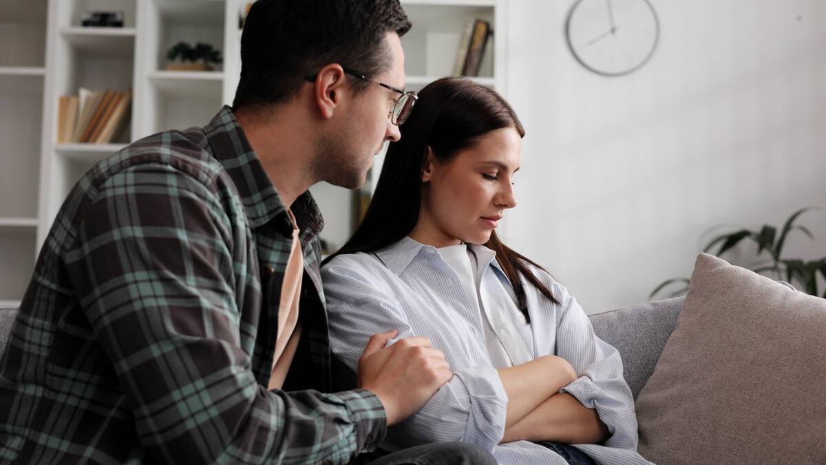 A man and woman sitting on a couch, the woman with her arms crossed looking upset, the man with one hand on the woman's arm, trying to apologize.