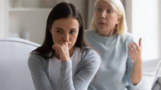 A woman sitting with her hand to her face, looking annoyed, while presumably her mother speaks with a terse expression behind her.