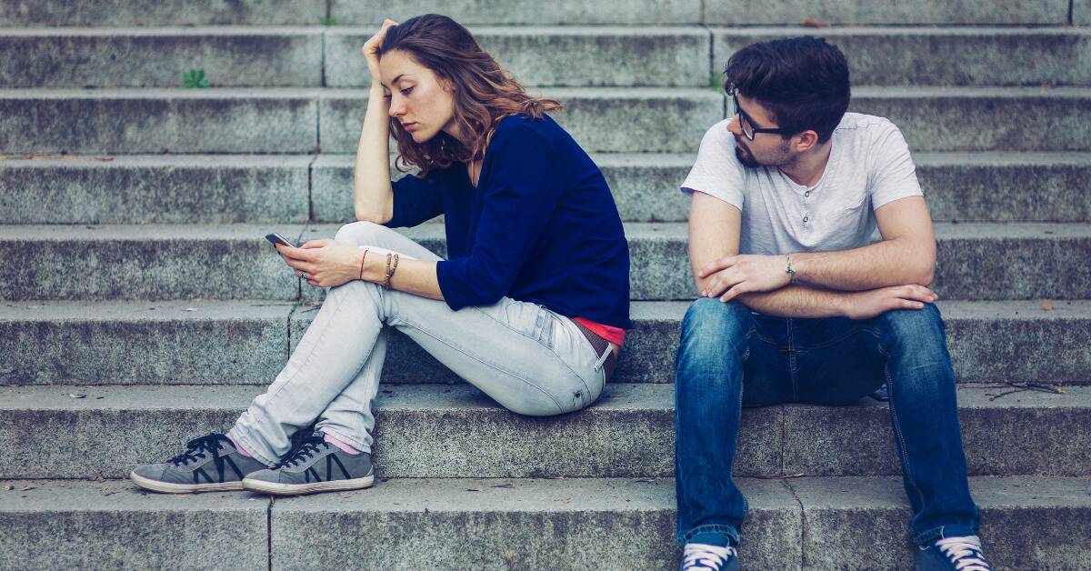 A man and a woman sitting on a set of stone stairs, the woman facing away from the man and looking at her phone.
