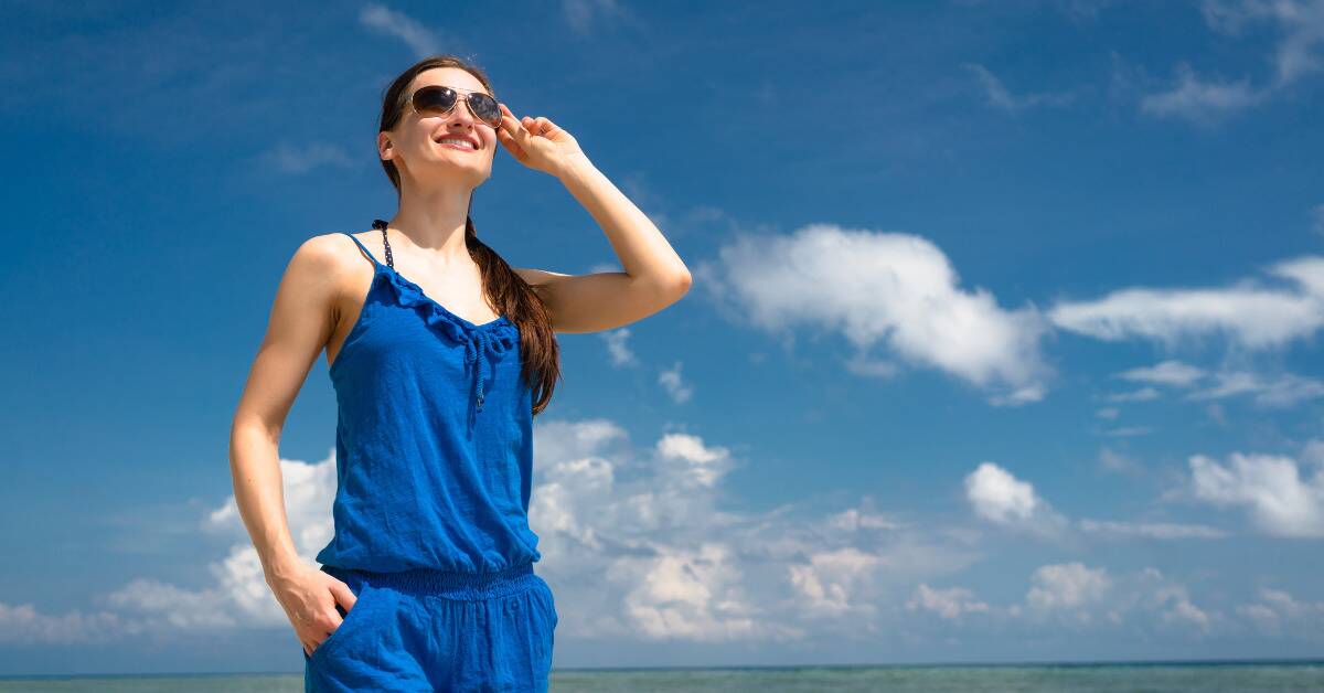 A woman standing outside confidently, one hand on her hip, the other adjusting her sunglasses, the bright blue sky her backdrop.