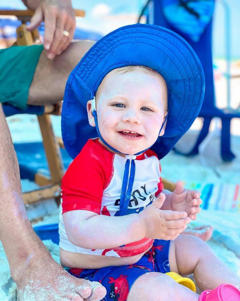 Beau sitting in the sand at the beach, sand on his hands and face, wearing a large blue sun hat.
