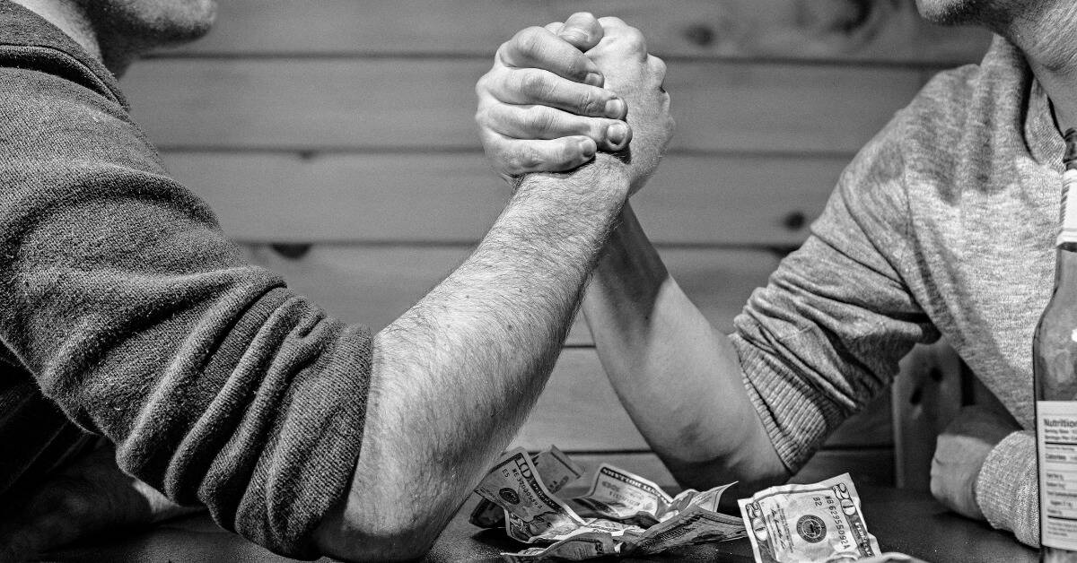 A greyscale image of two men about to arm wrestle, cash bets thrown on the table below them.