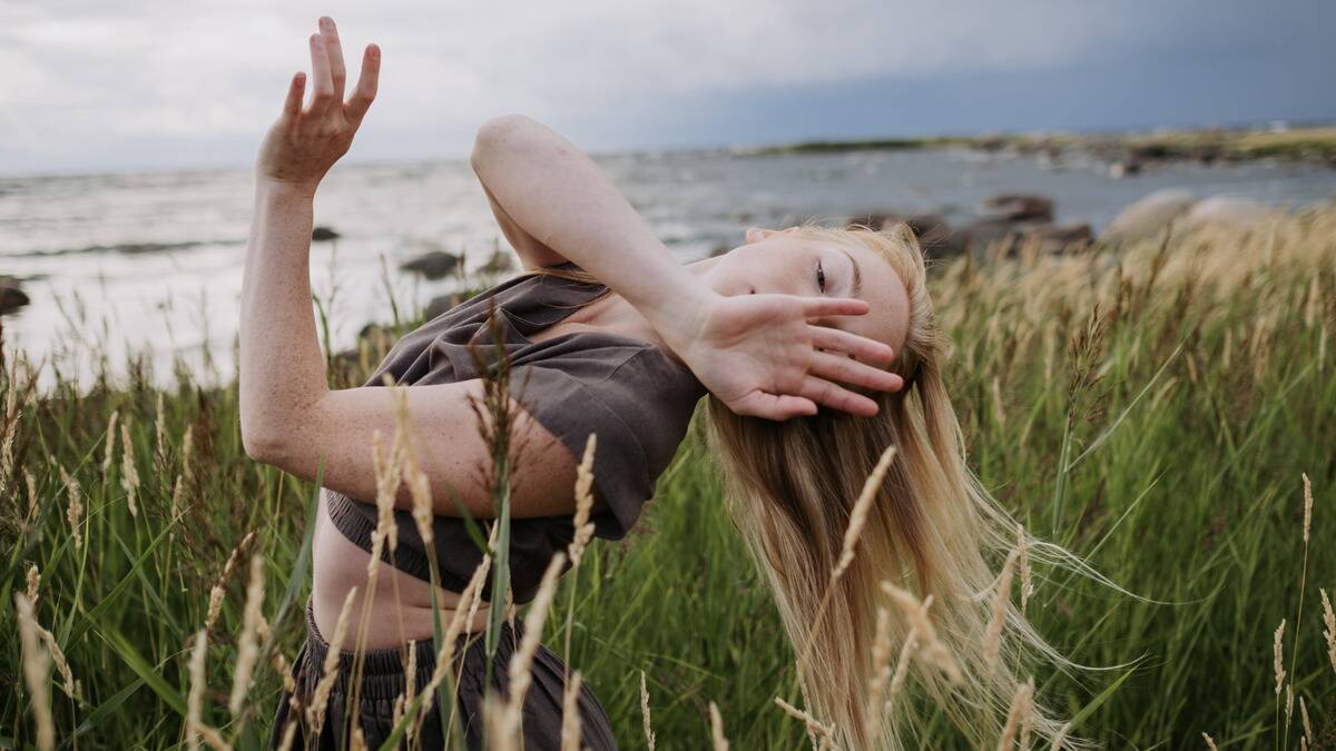 A woman dancing in a field, leaning backward and making flowing movements with her arms, her face covered by her hand.