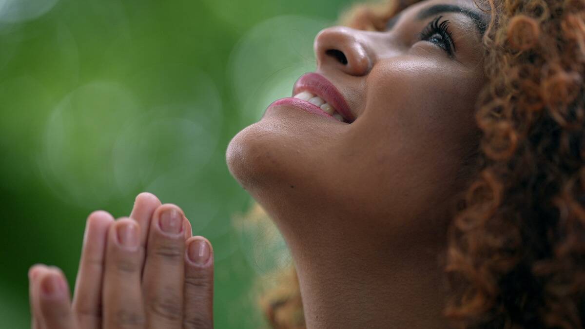 A close shot of a woman looking up to the sky as she smiles, her hands together in a prayer motion, though only the top halves of her fingers are visible.