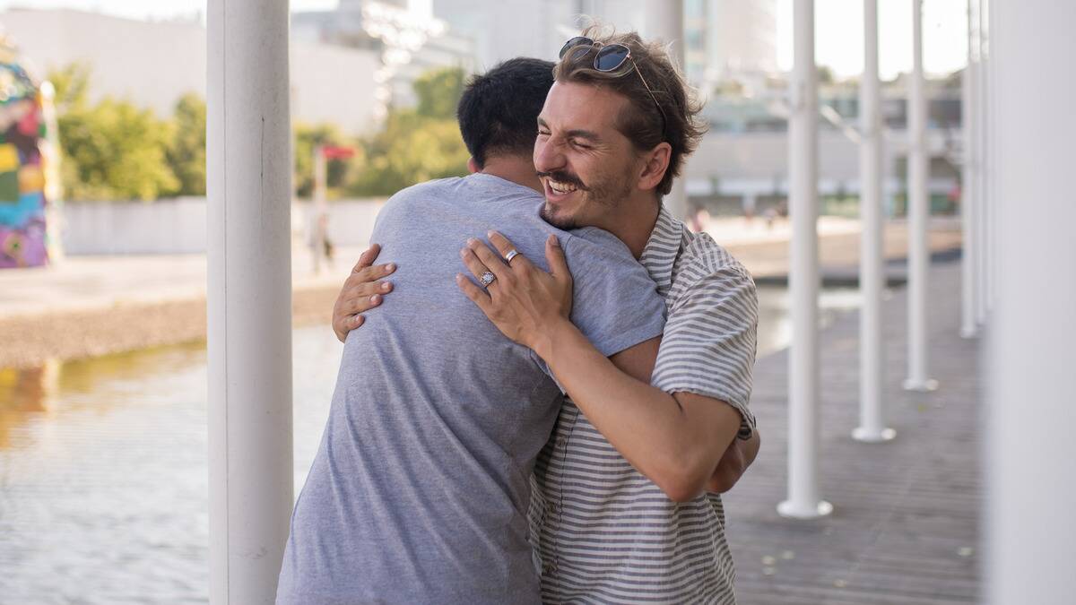 Two friends hugging outside on a boardwalk. The man whose face we can see is smiling brightly with his eyes closed.