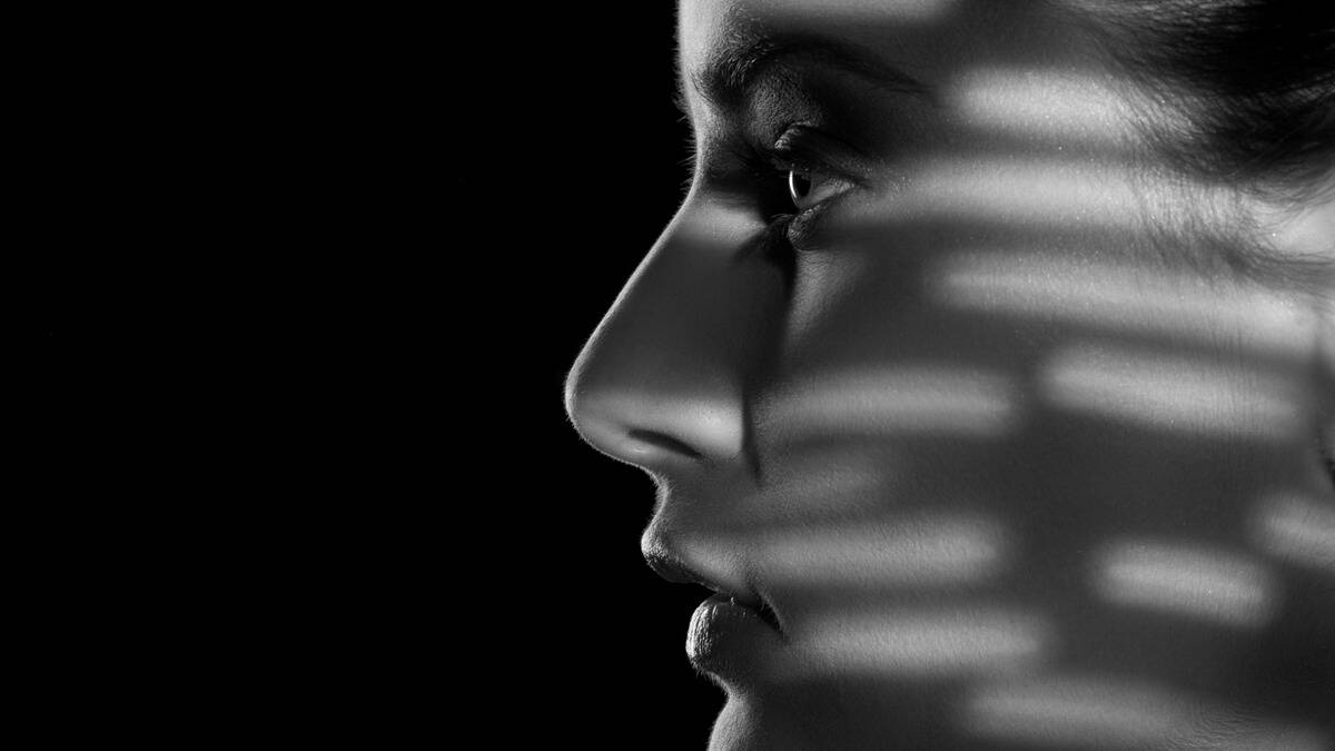 A close, greyscale image of a woman's face in profile, shadowed but being lit but small bars of light.