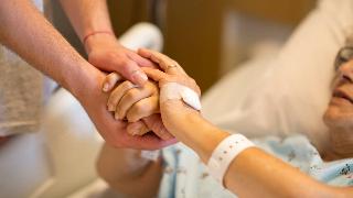 Someone using both hands to hole the two hands of a woman in a hospital bed, a bandage over her hand.