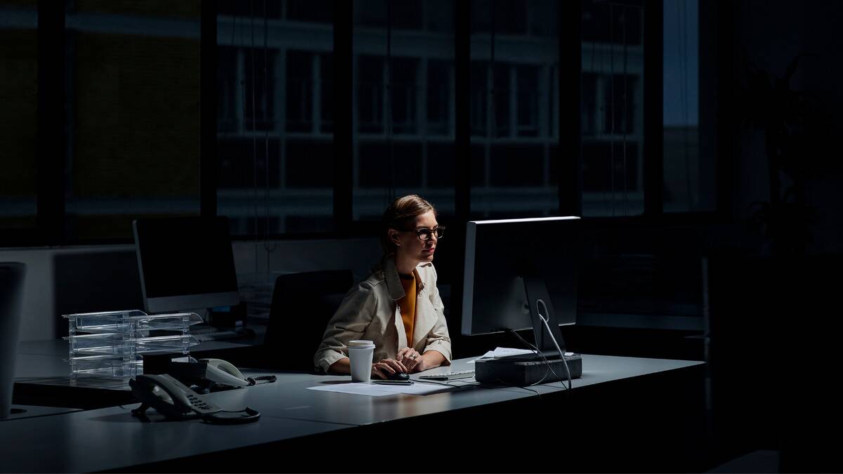 A woman sitting at her desk in an office working on something, the sky dark behind her, indicating how late she's working.