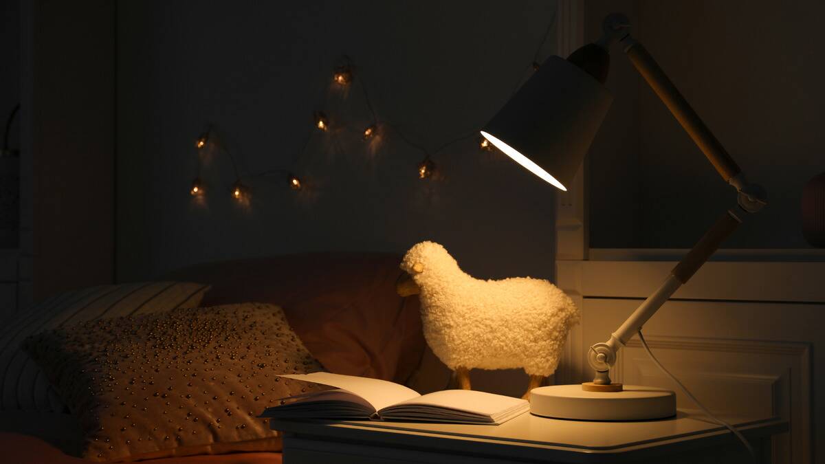 A lamp on a nightstand in a child's bedroom, a plush sheep and a book open beside it.