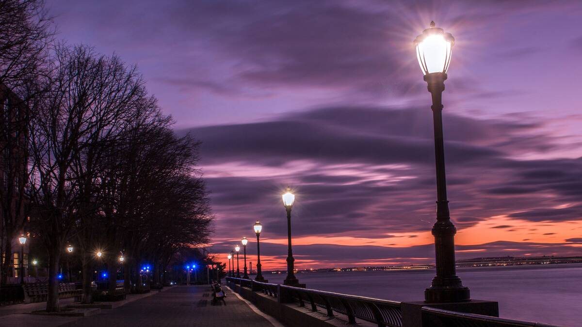 A path next to some water lined by streetlights, a purple sunset sky in the background.