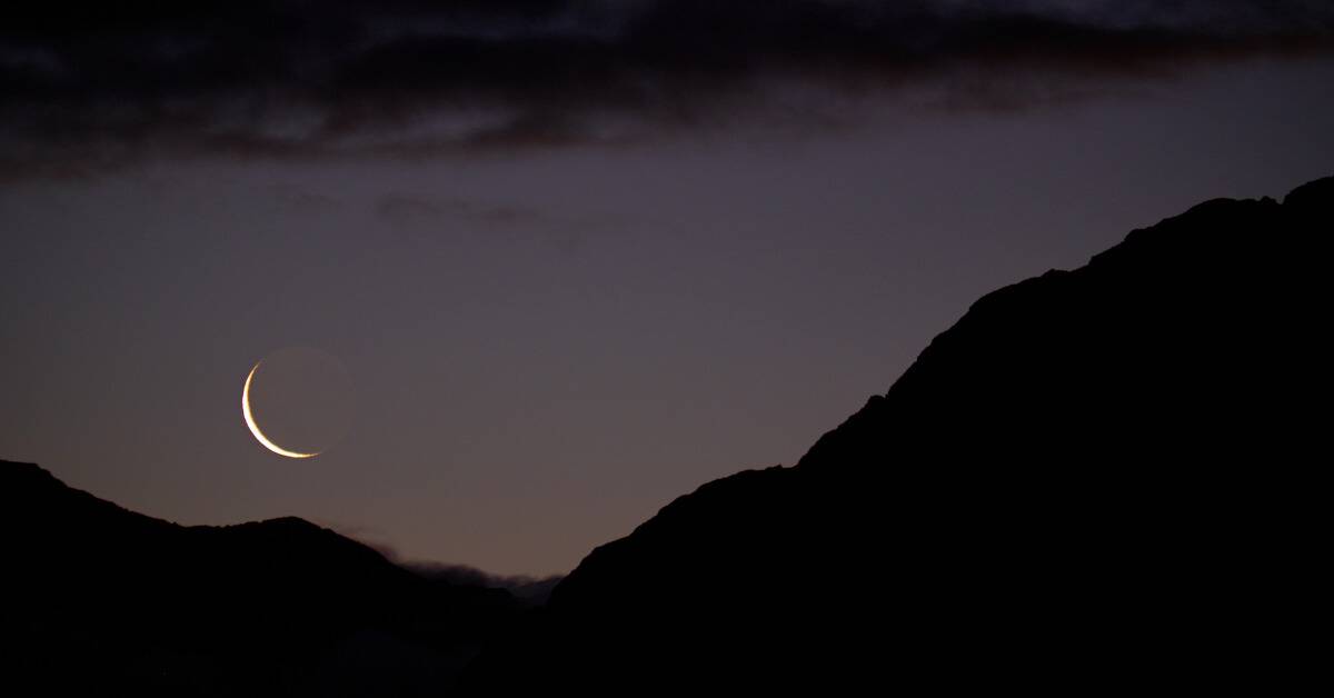 A crescented new moon hanging low in a dusky sky.