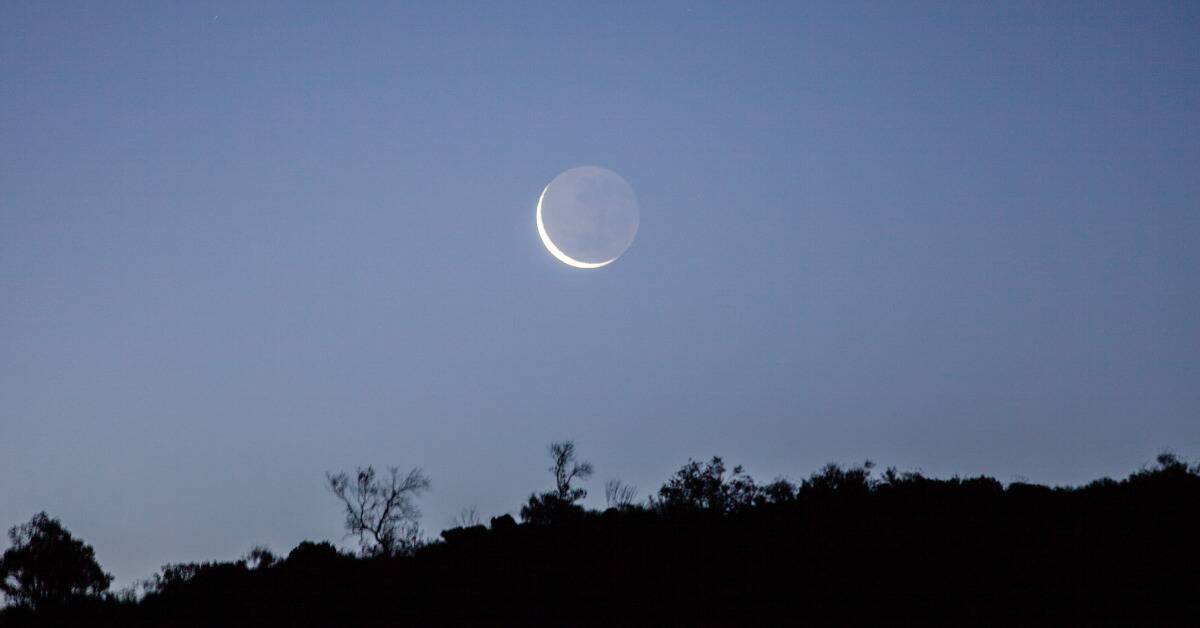 A crescented new moon visible in the blue dusk sky above a shadowed hill range.