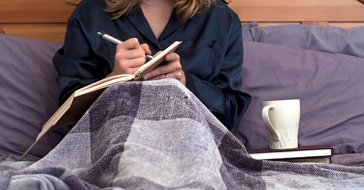 A close shot of a woman sitting with her knees drawn up under a blanket, writing in a journal.