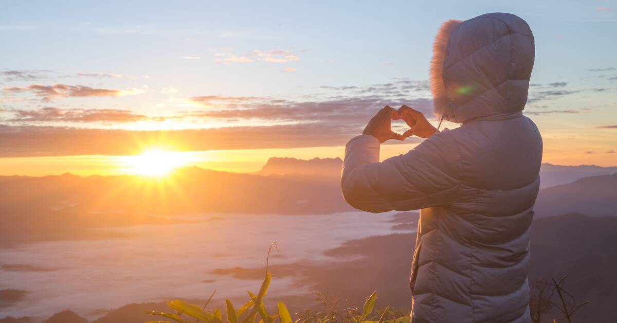 Someone in a puffy coat standing high on a hill range, making a heart with their hands toward the rising sun.