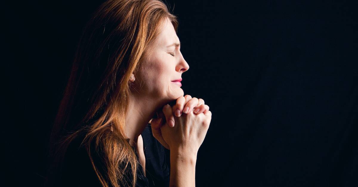 A woman with her hands together in prayer, eyes closed, against a black background.