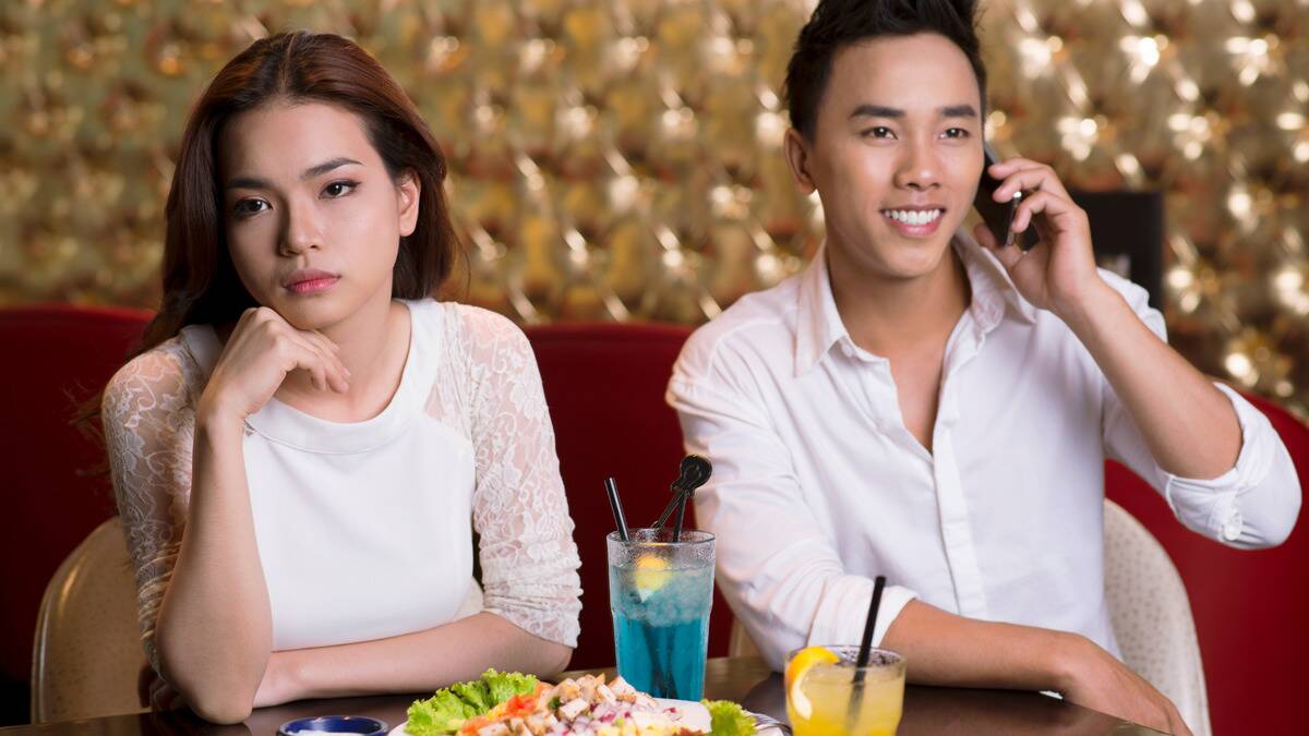 A couple out for dinner, sitting side by side, the man talking on the phone while the woman looks bored.