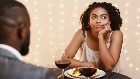 A woman sitting across from a man at dinner, her cheek in her hand, looking bored and annoyed.