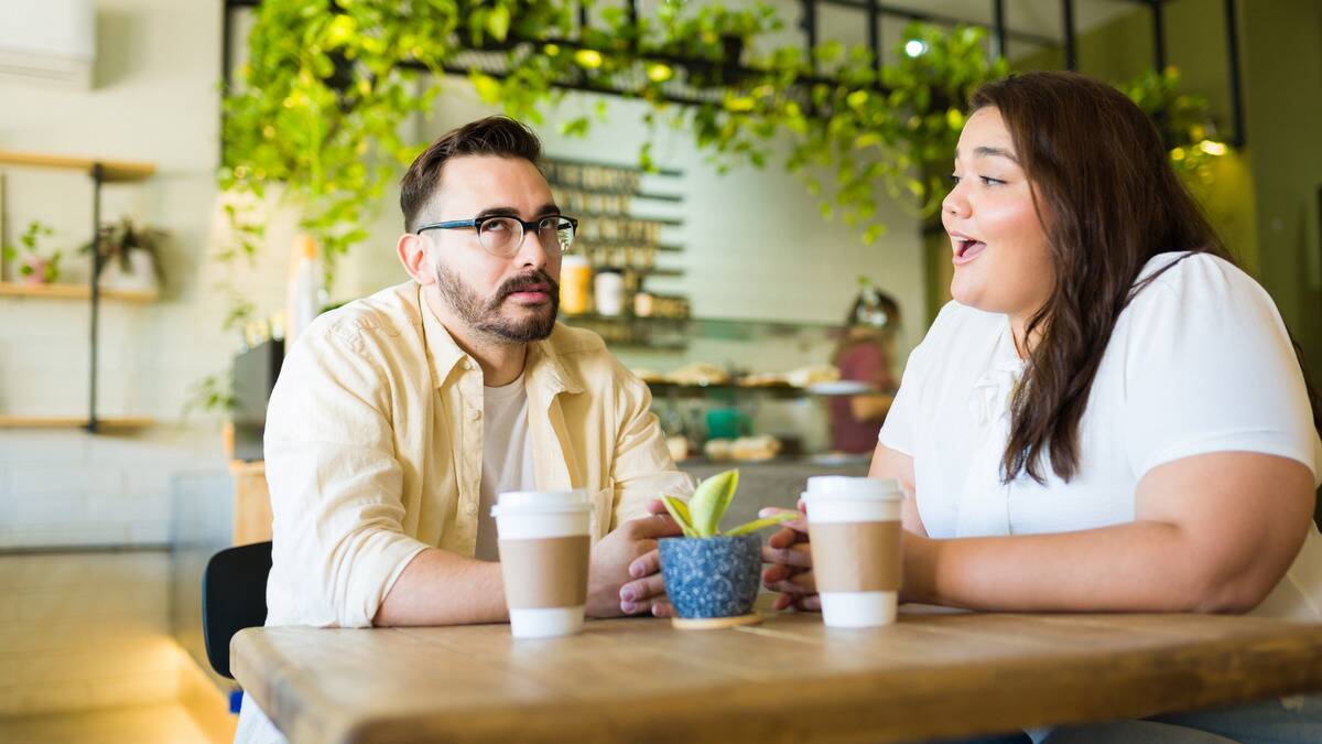 A man rolling his eyes as a woman speaks to him, both sitting in a cafe.