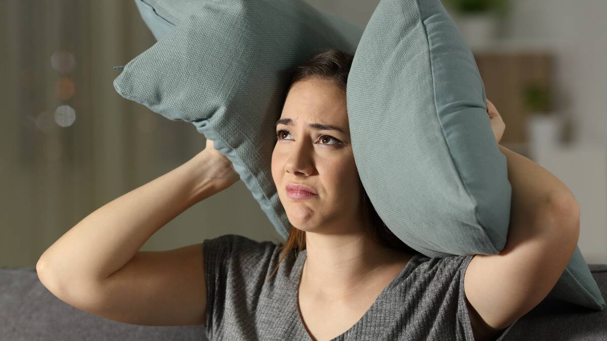 A woman looking annoyed and frustrated, holding pillows up against her ears.