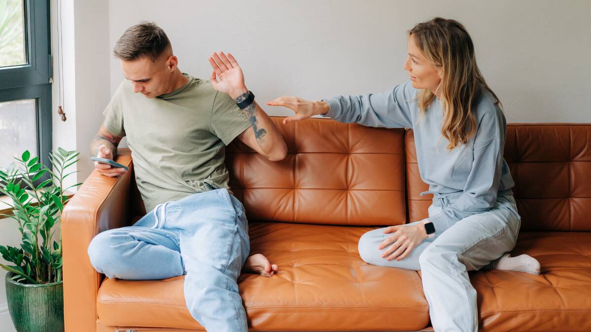 A couple sitting on a couch, the man looking at his phone and waving away the hand of his girlfriend who's trying to reach out to him.