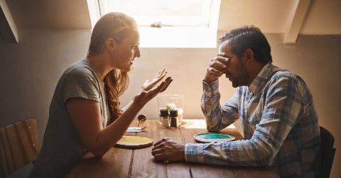 A couple seated across a table from one another, the woman speaking critically and the man with his face in his hand.