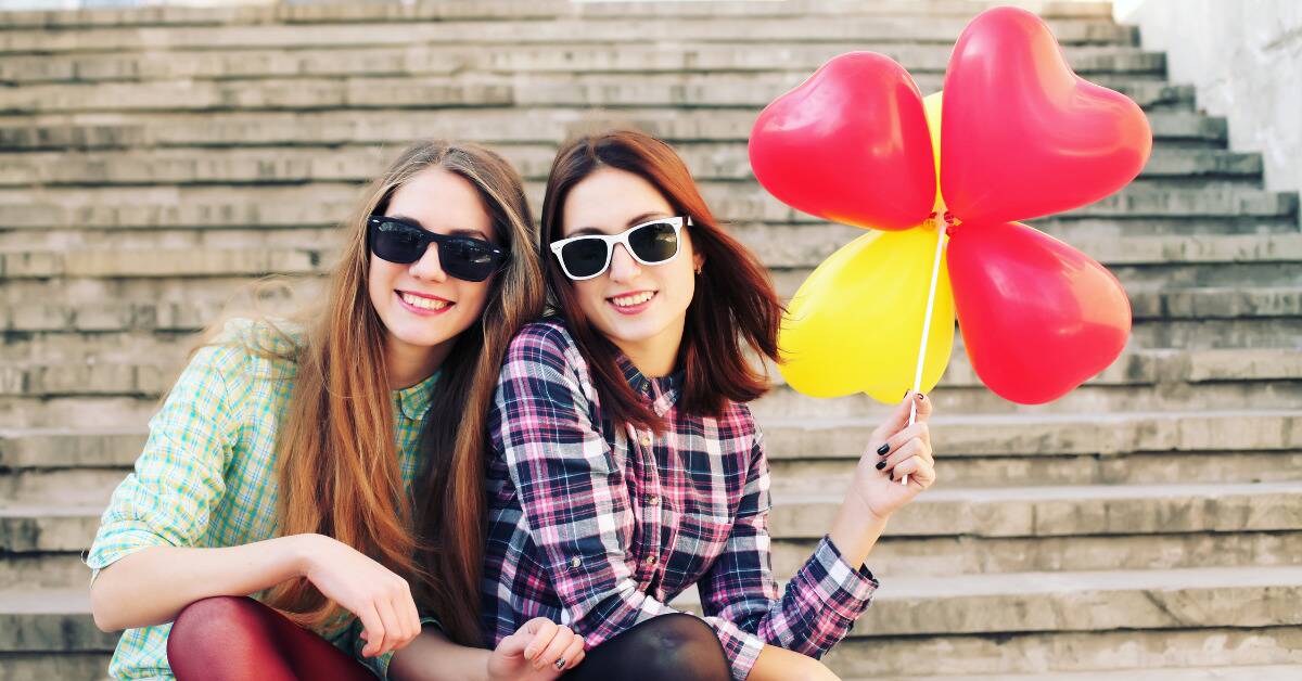 Two sisters in sunglasses sitting on some outdoor steps, one holding a bouquet of heart-shaped balloons.