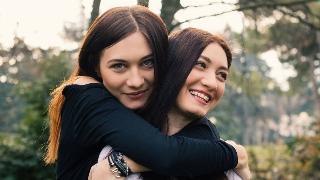 Two sisters hugging, one hugging the other from behind, both smiling.