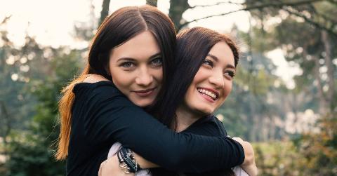 Two sisters hugging, one hugging the other from behind, both smiling.
