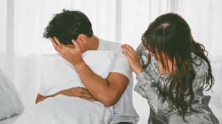 A man hugging a pillow and covering his face, his girlfriend placing a comforting hand on his shoulder as they both sit in bed.