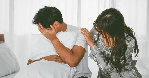 A man hugging a pillow and covering his face, his girlfriend placing a comforting hand on his shoulder as they both sit in bed.