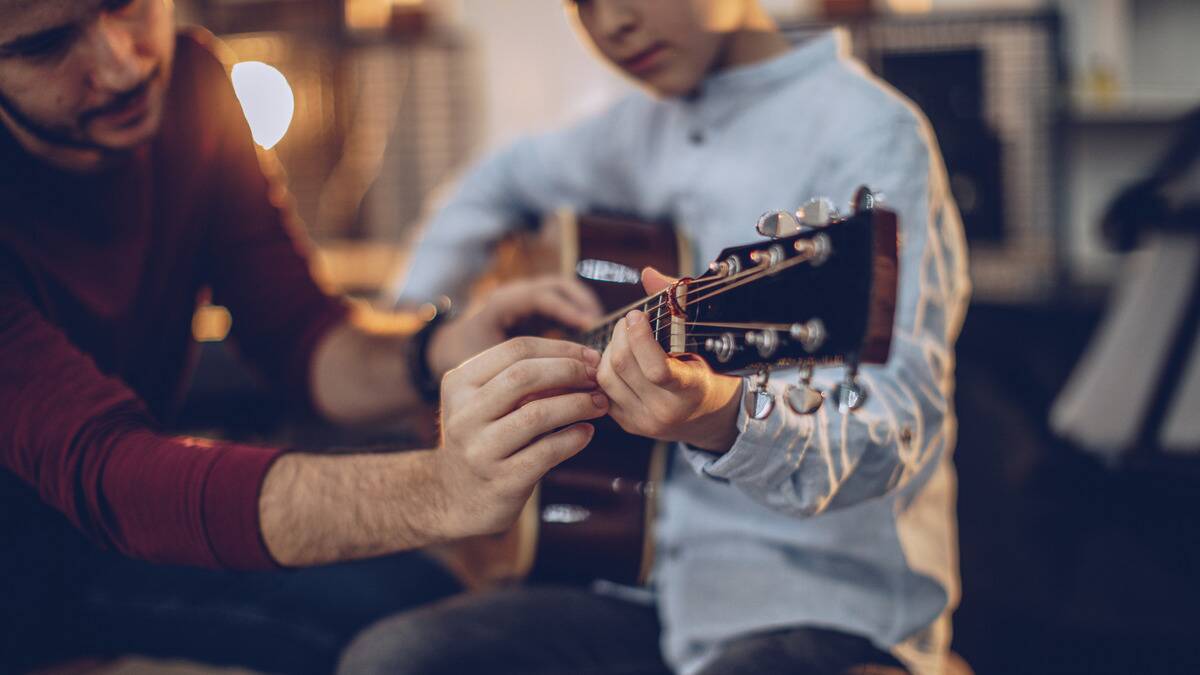 A close shot of an instructor telling a young boy how to position his hands on the neck of a guitar.