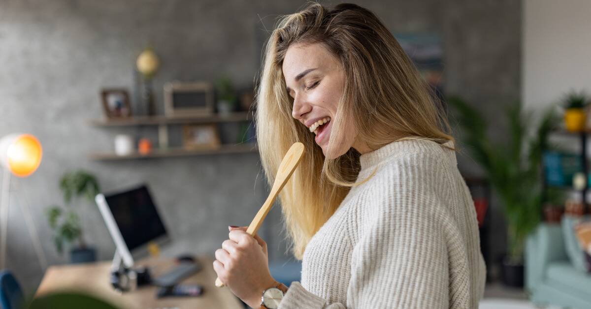A woman singing into a wooden spoon as if it were a microphone.
