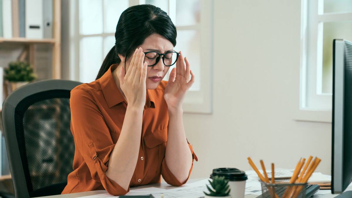 A woman sitting at her desk, hands on the frames of her glasses, looking frustrated.