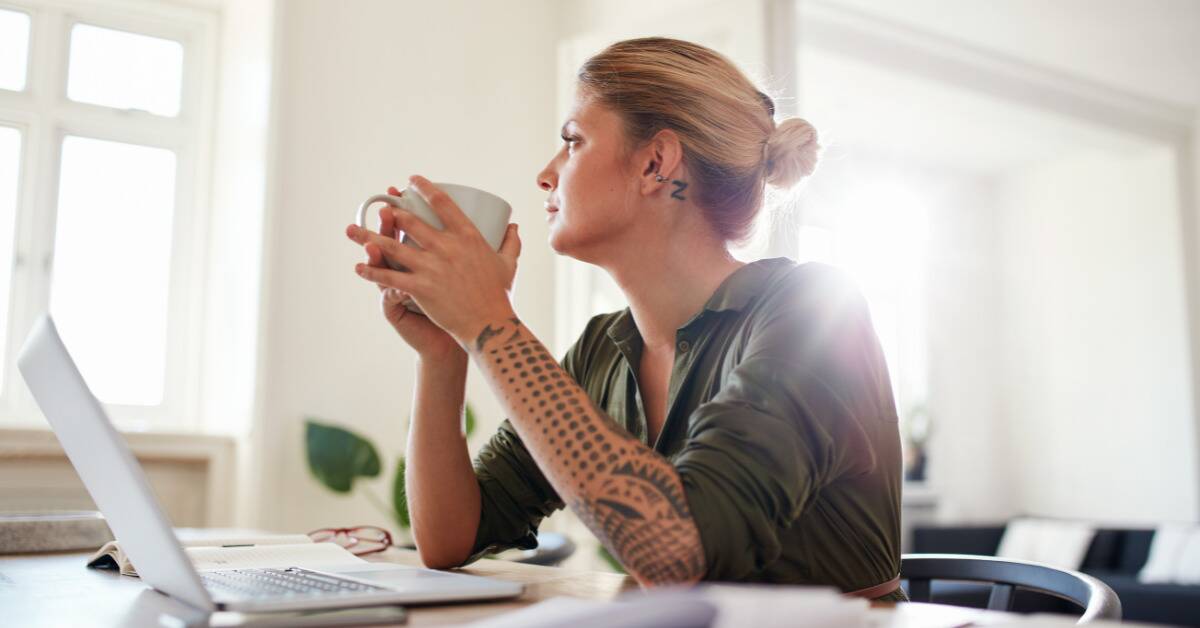 A woman sat at her desk holding a mug in both hands, looking out her window whistfully.