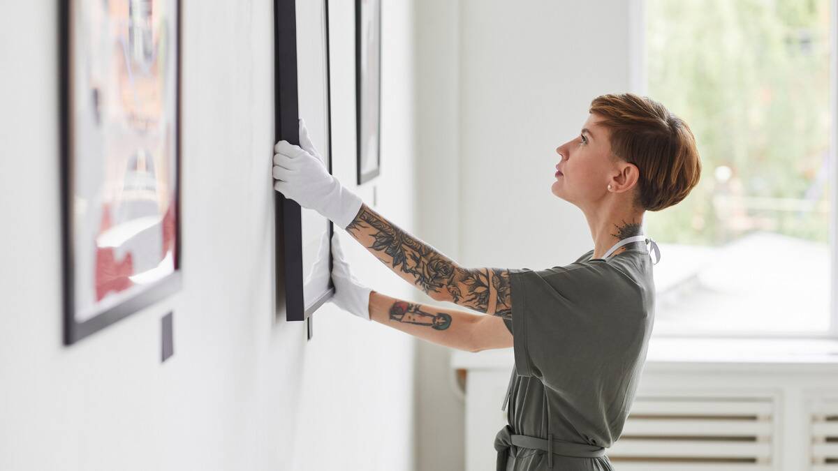 A woman hanging up a piece of art on a wall, wearing gloves so she doesn't damage it.