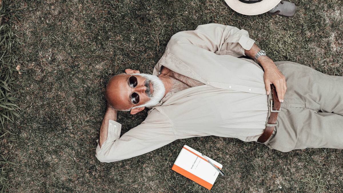 A man laying down in the grass, sunglasses on, one hand behind his head, a book and a hat off to his side.